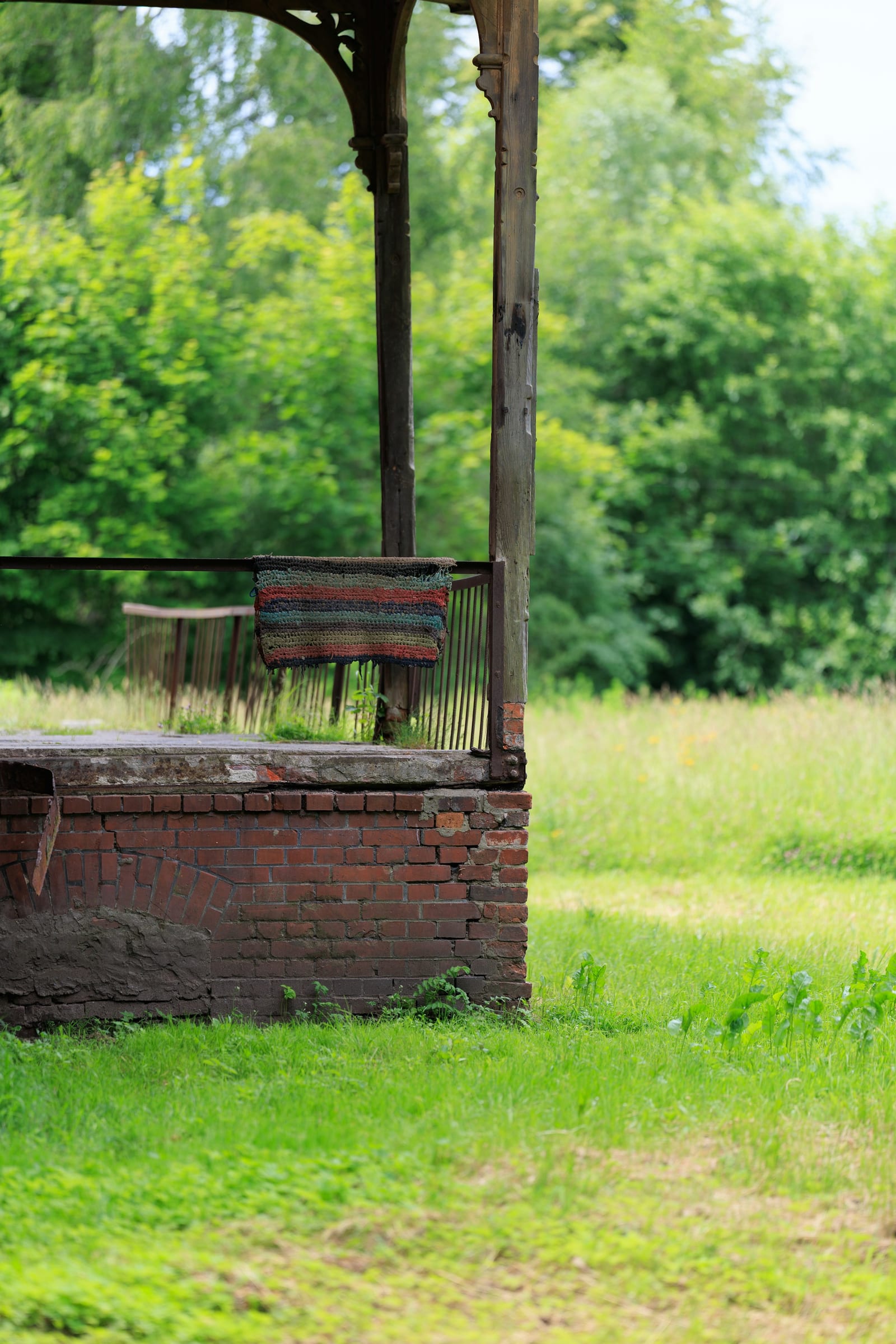 A weathered brick-and-wood porch with a worn rug over the rail, overlooking a green field.