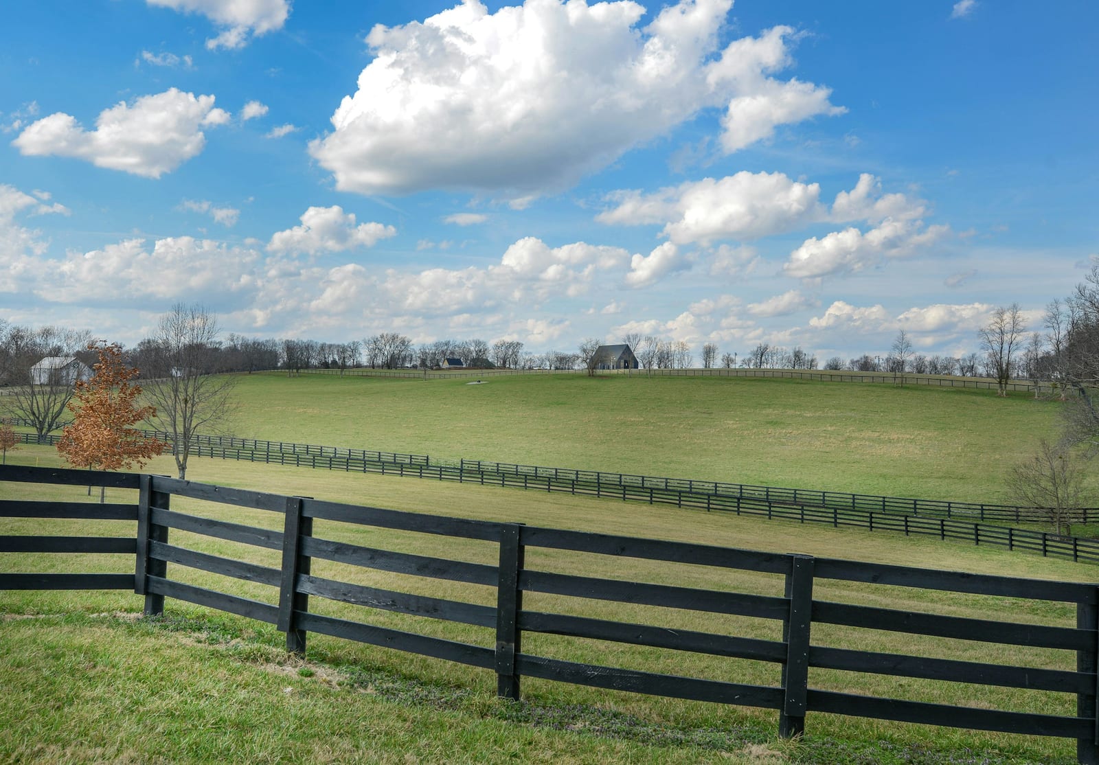 A black four-plank fence running along rolling green Kentucky pasture under a blue sky with scattered clouds.